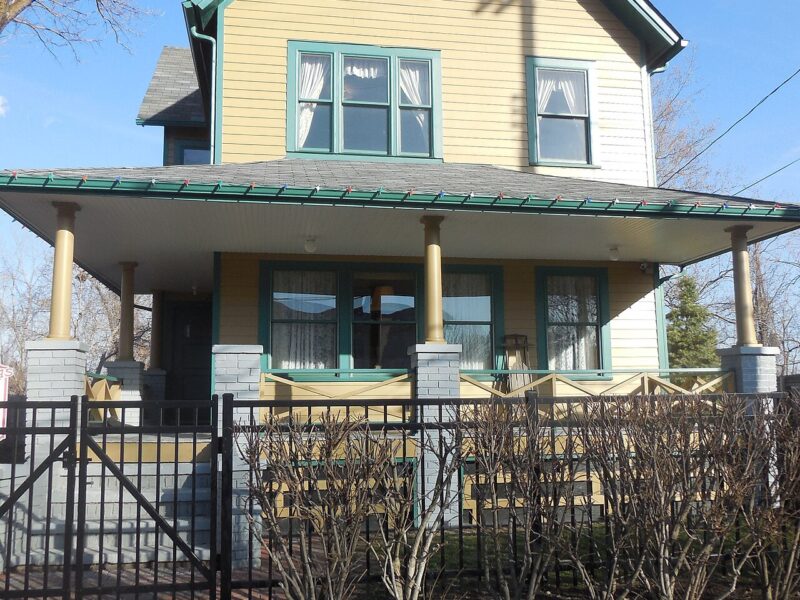 Yellow two-story house with green trim and a wide front porch behind a black iron fence in Cleveland, Ohio
